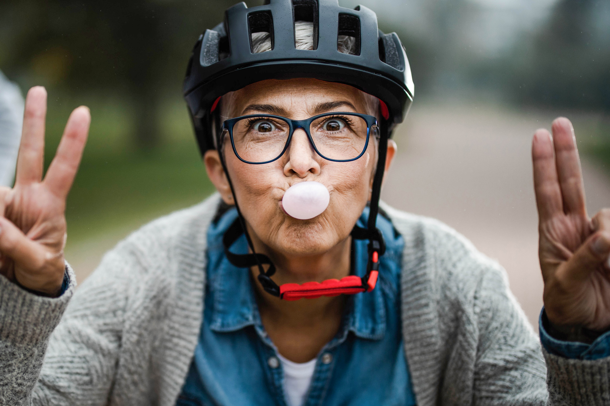 A playful senior woman shows peace signs while blowing bubble gum in a park.