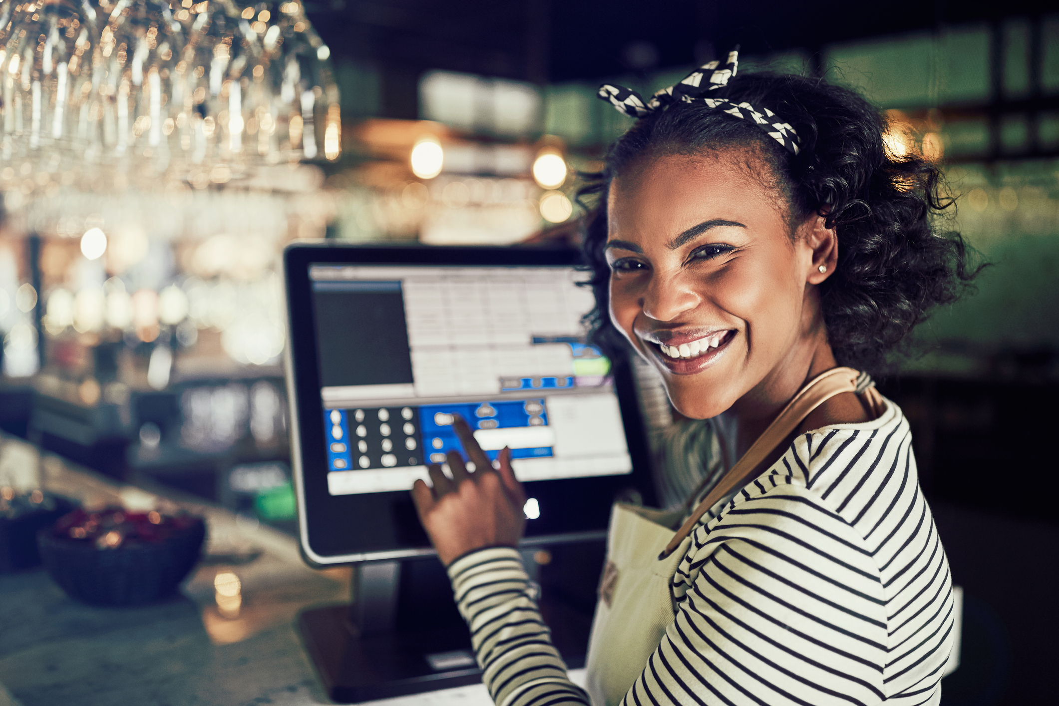 A smiling young retail associate using a touchscreen point-of-sale monitor at the checkout counter.