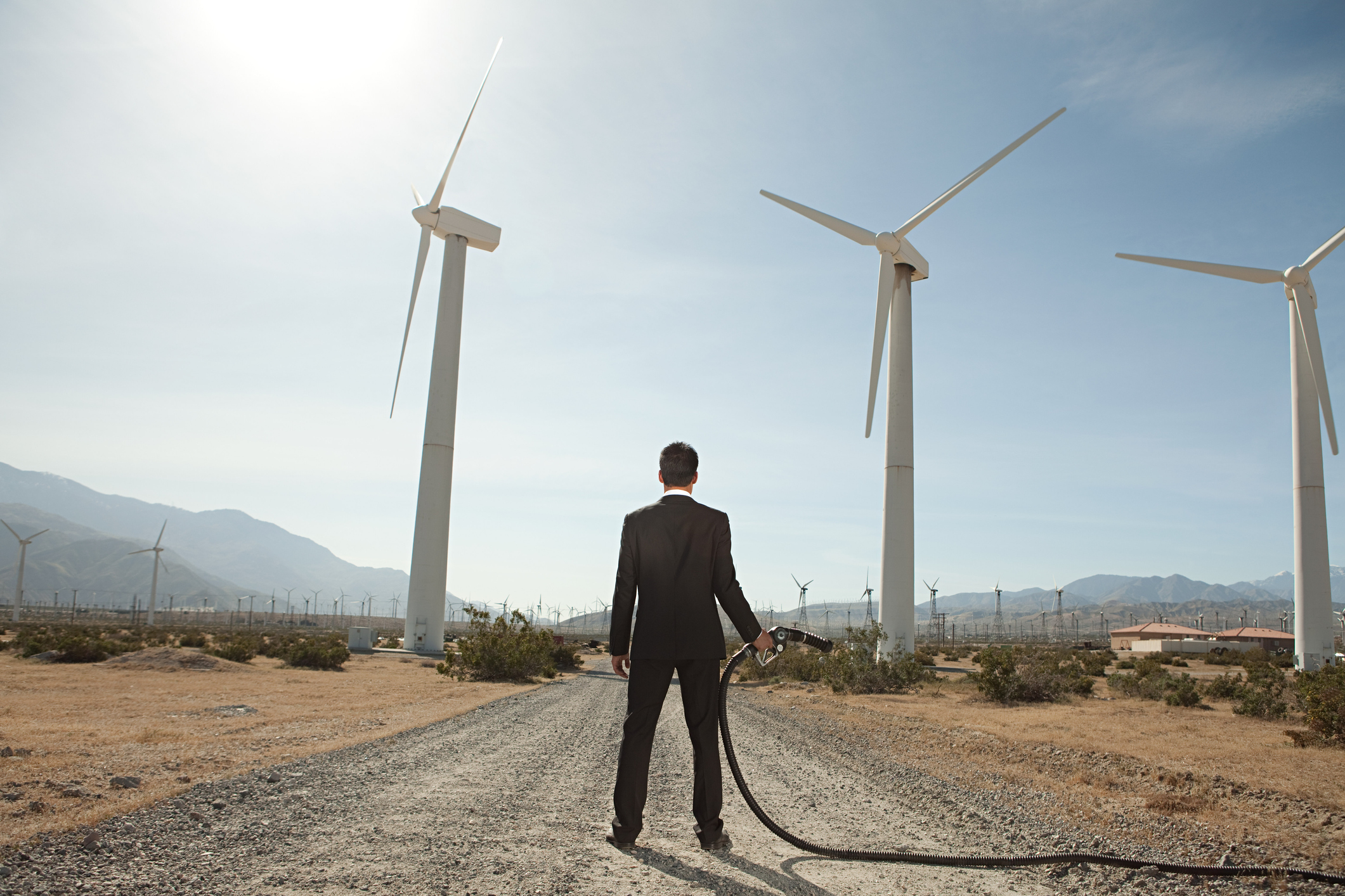 A man standing in the middle of a road holding a gas pump while gazing at a field of wind turbines.
