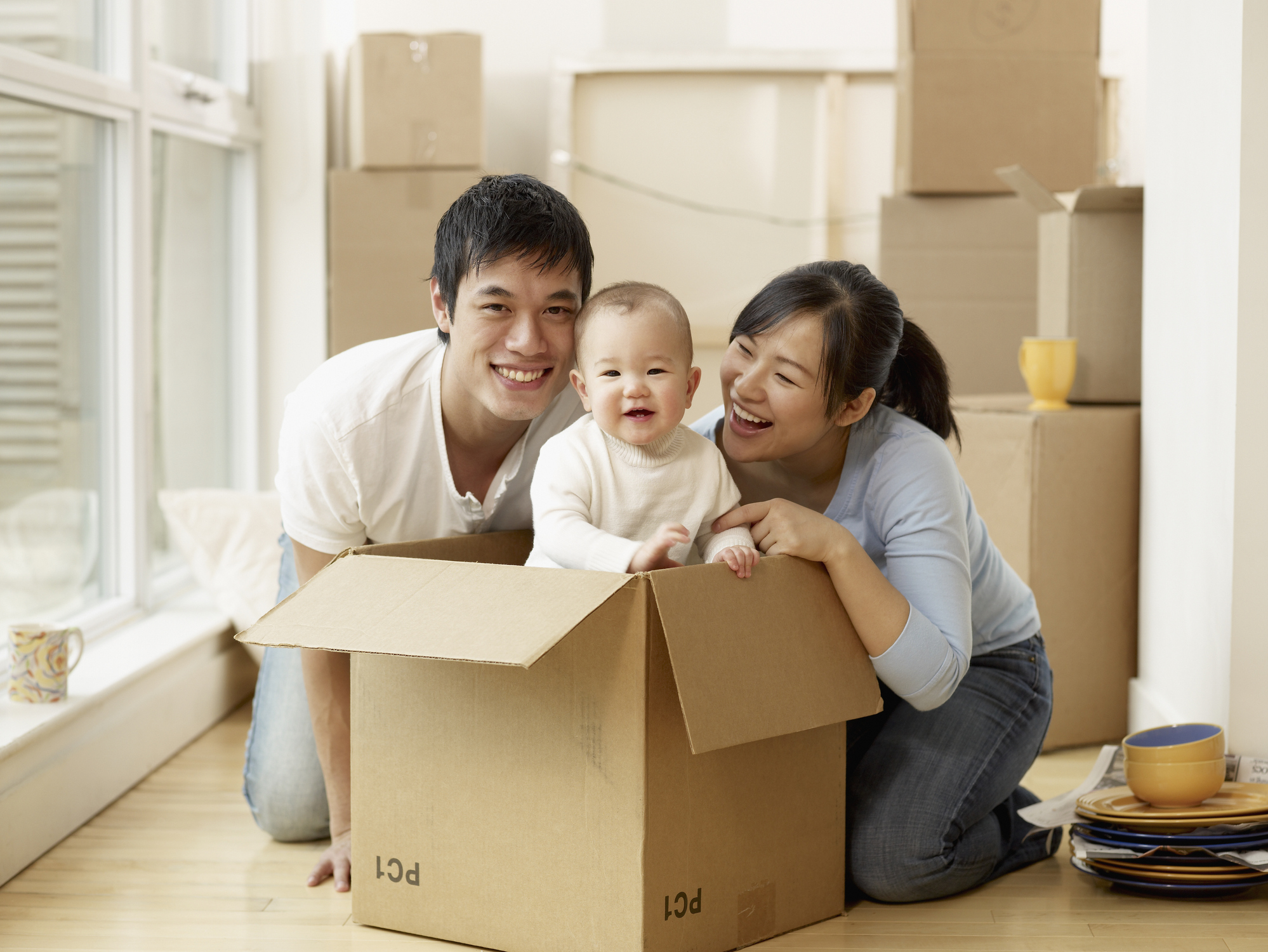 An Asian family laughs about an infant standing in a moving box as other boxes sit behind it in a home.