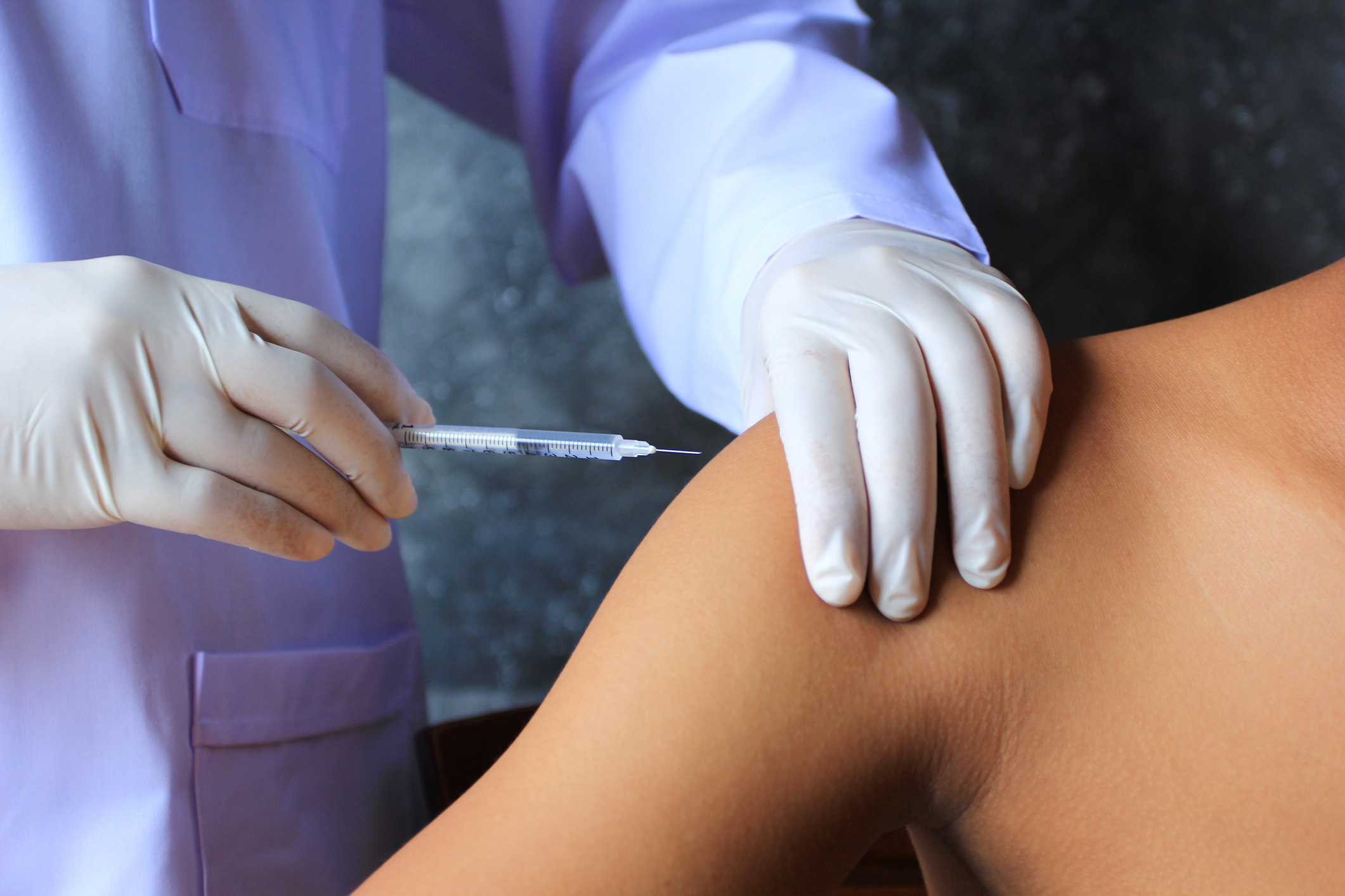 Gloved hands giving an injection into someone's shoulder.