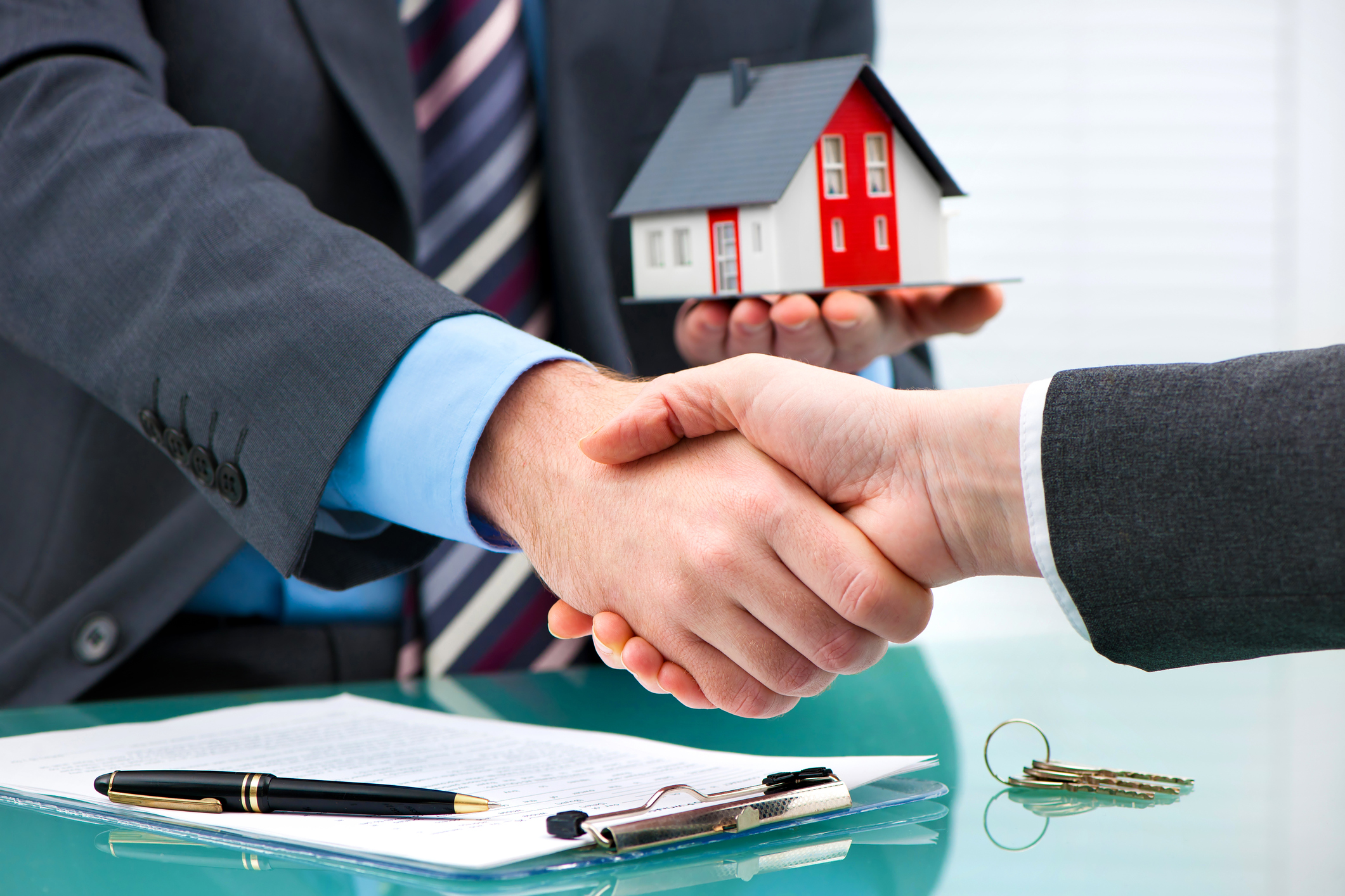 Two businessmen in suits shaking hands, with one holding a miniature house in his left hand.