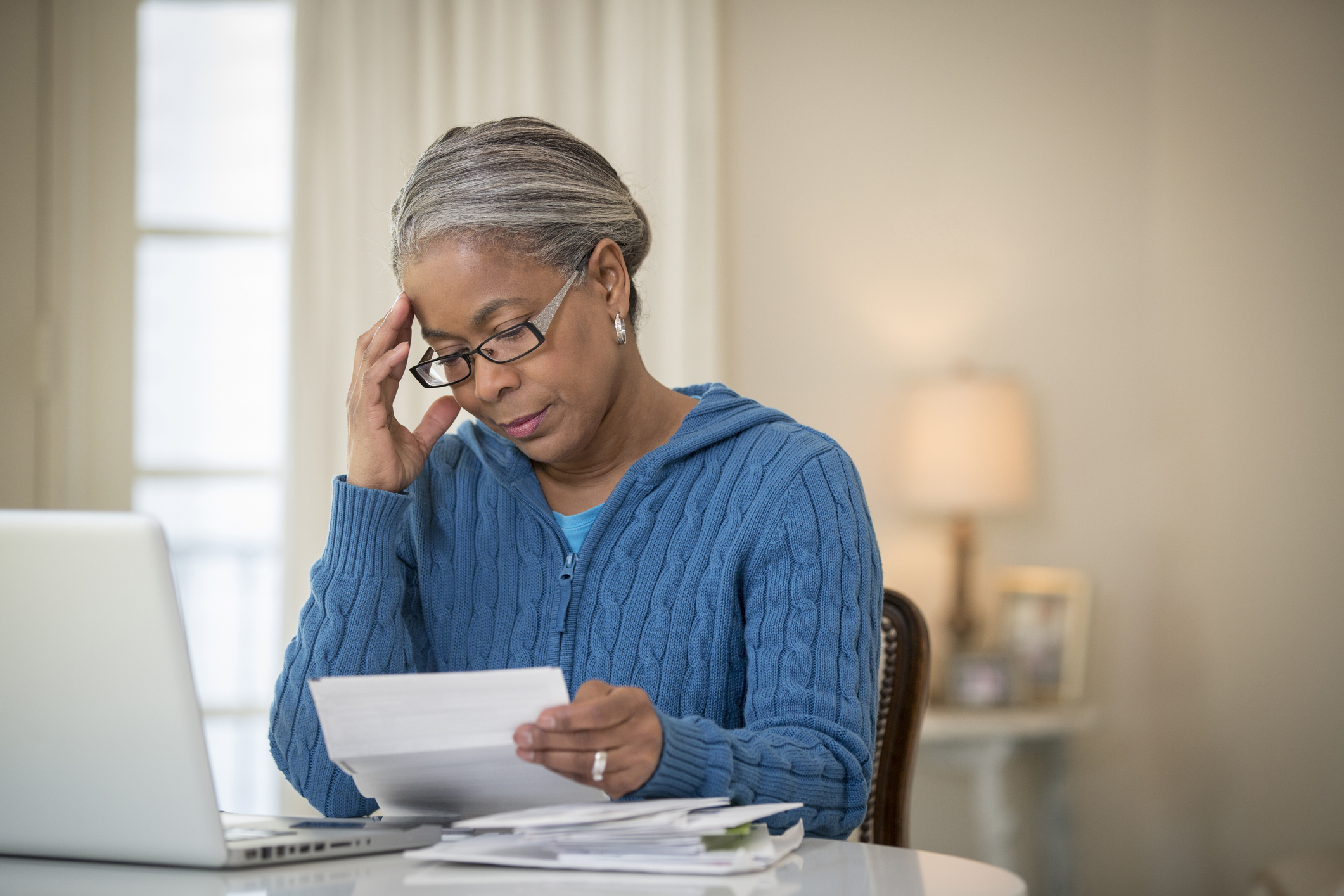 Senior woman wearing blue sweater sits in front of a laptop, glancing over bills.