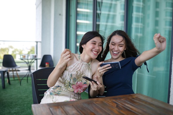 Two young girls clenching celebratory fists while playing with a smartphone on a balcony. 