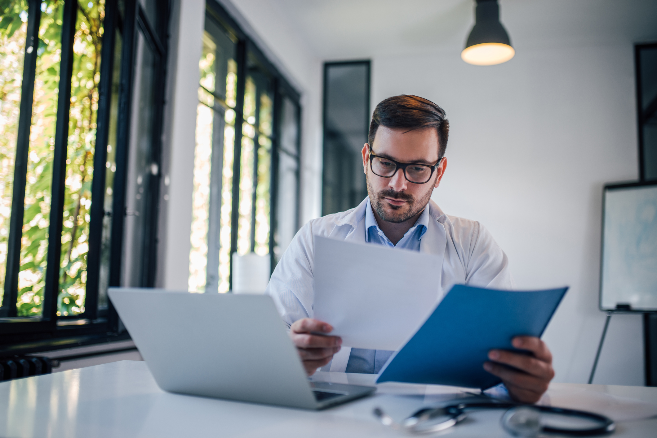 A doctor sitting at a table by a window studies a sheet of data.