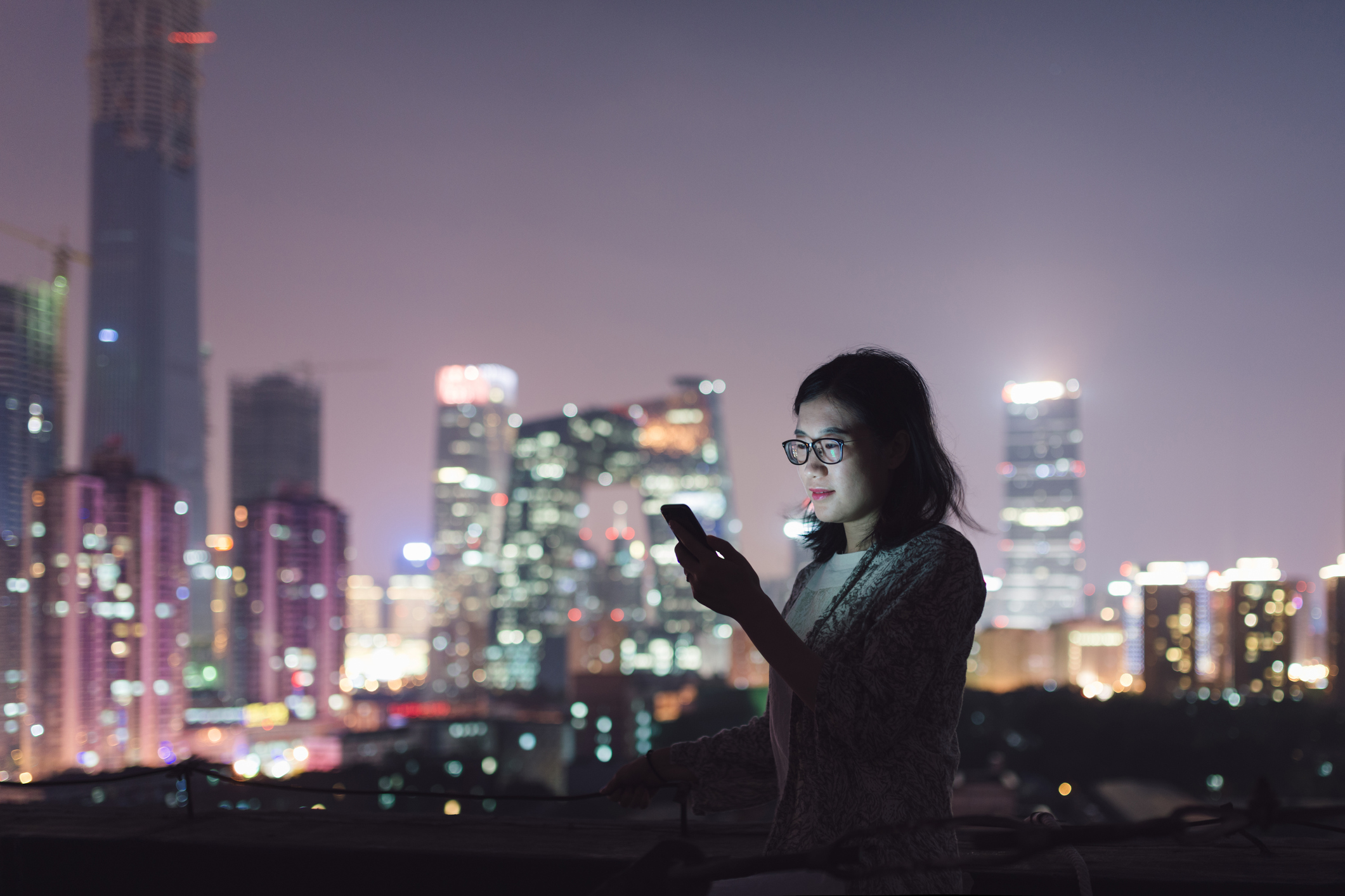 Young woman in glasses looks at her phone  in front  of a cityscape at night.