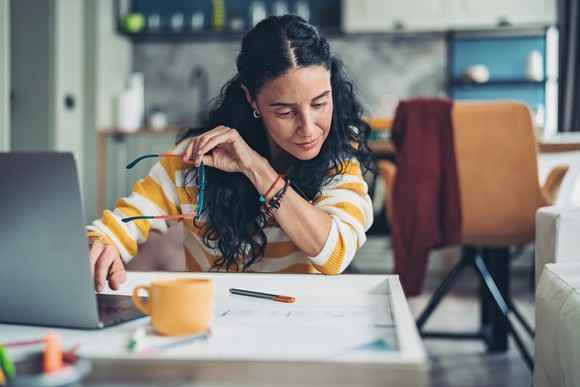 Woman looks at a piece of paper on her desk while sitting in front of a mug and her laptop.