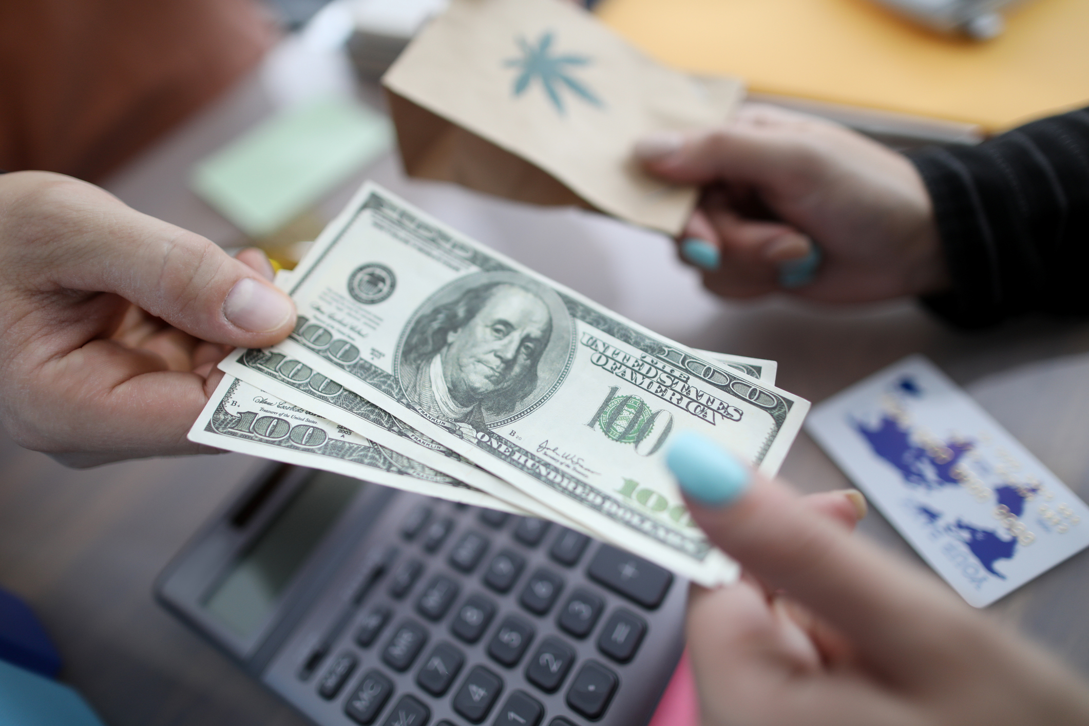 Closeup on hands exchanging dollar bills for a bag with a cannabis leaf printed on it.