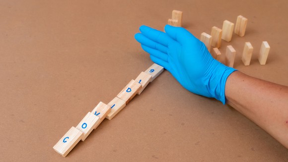 Gloved hand between wood dominos spelling COVID-19 that have fallen down and other wood dominos that are standing up