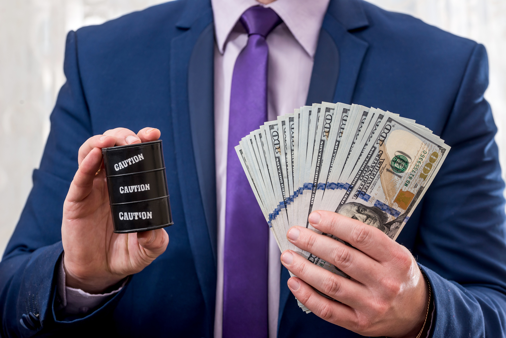 A man holding a barrel of oil with caution written on it in one hand and cash in the other hand.