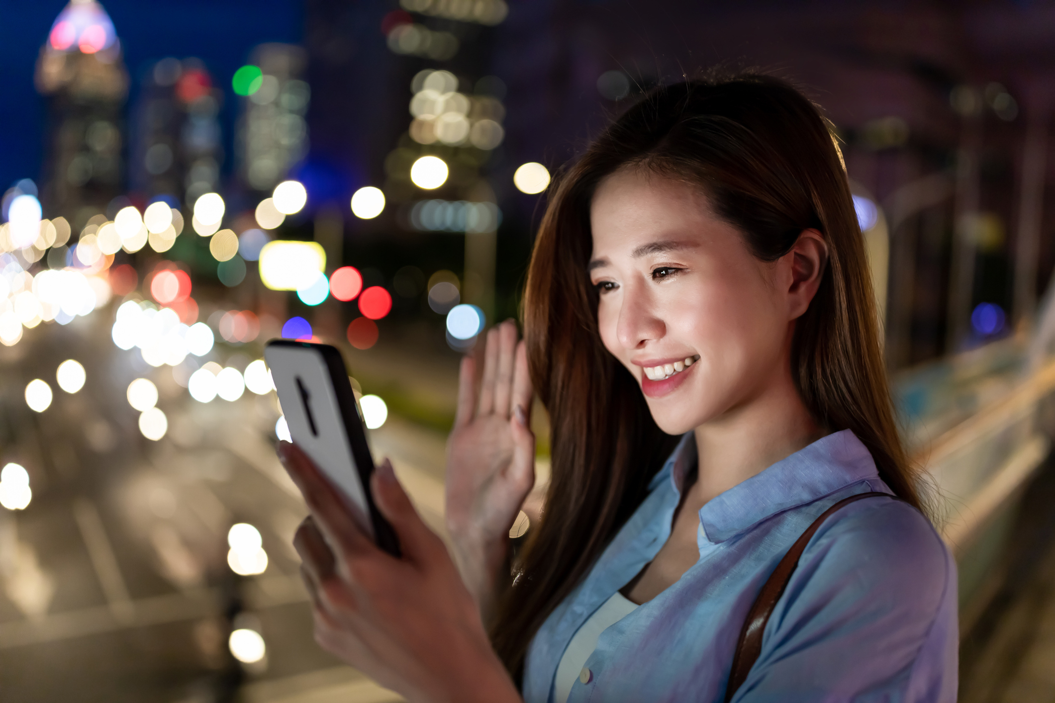 Young woman smiling as she waves to someone on her smartphone while walking in the city.