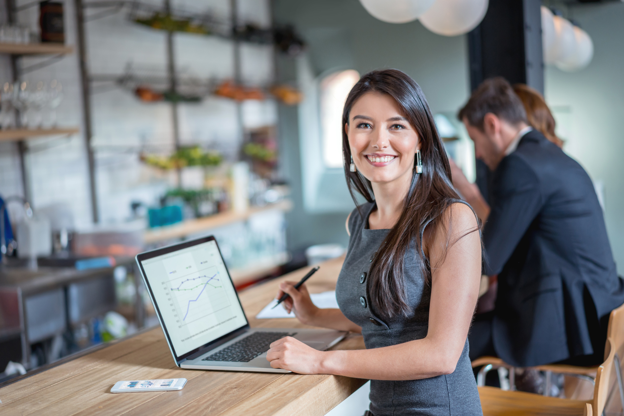 Young adult stands at a bartop with her laptop and pen in hand.