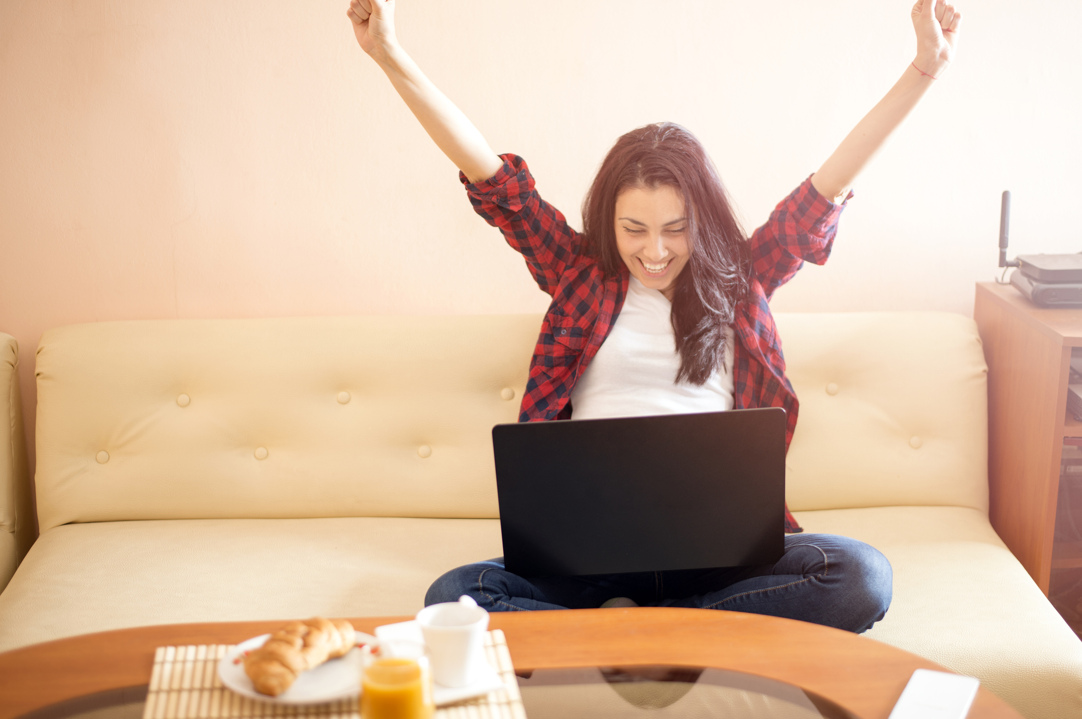 A young woman with a laptop on her couch, lifting her hands over her head with a big smile.