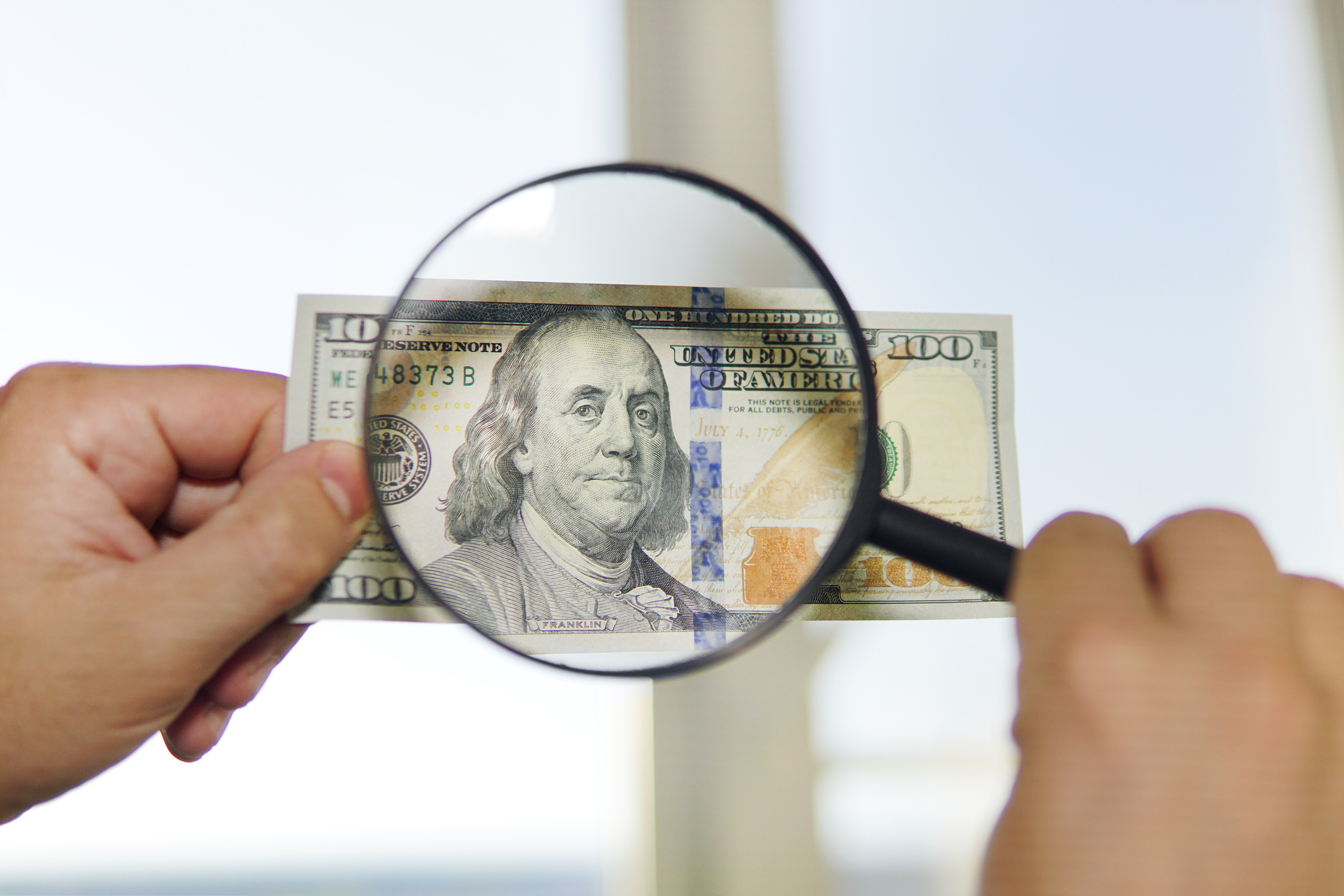 A person examines a one hundred dollar bill using a magnifying glass.