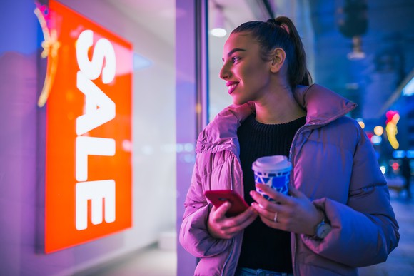 Woman holding a cup and a phone standing in front of retail shop.