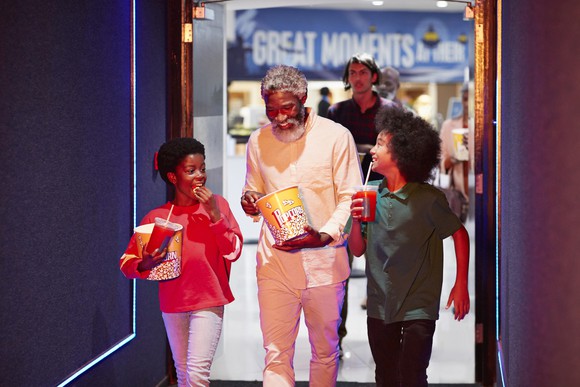 An adult with two kids walking into a movie theater with popcorn and drinks.