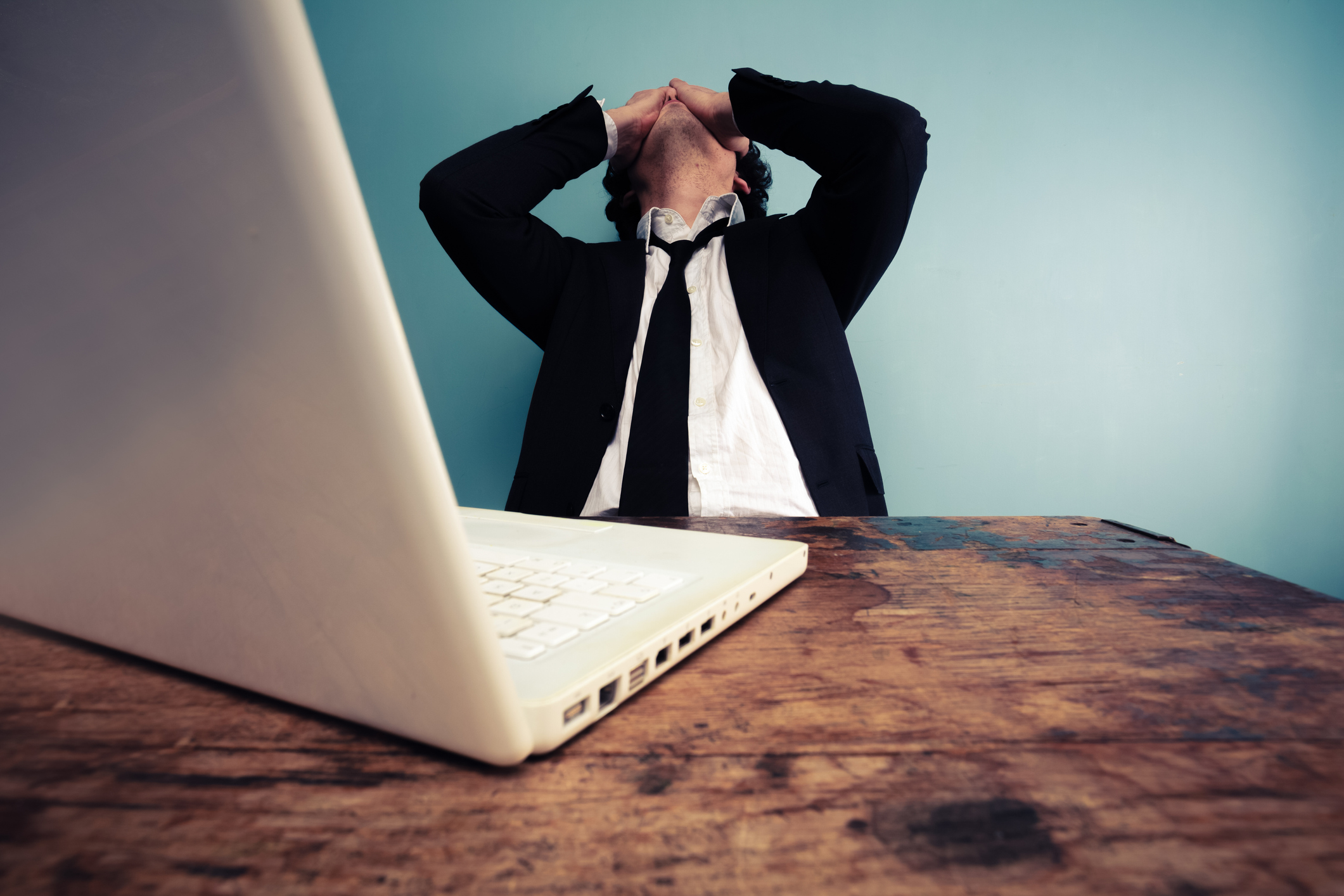 A man sits in front of computer. He is leaning back, with both of his hands covering his eyes.