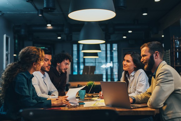 A group of five people meets around a desk with laptops and papers in front of them in an office.