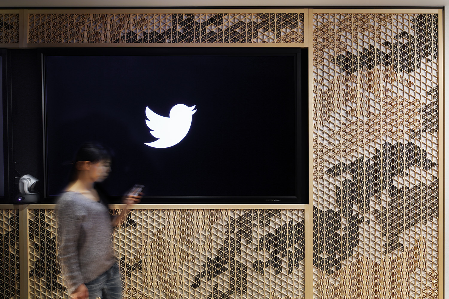 Person looking at their phone while walking by the Twitter logo displayed on a wall.