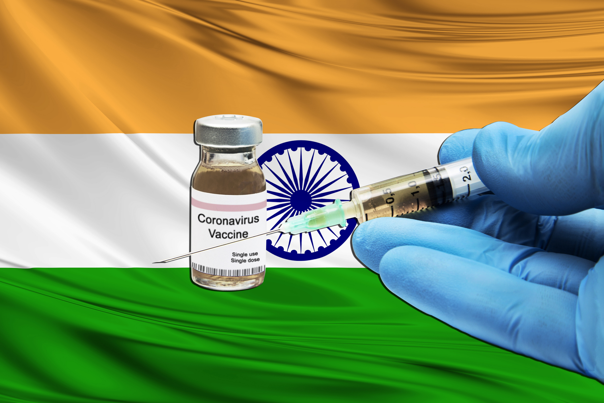 Gloved hands holding a syringe with needle next to a coronavirus vaccine vial with an Indian flag in the background.