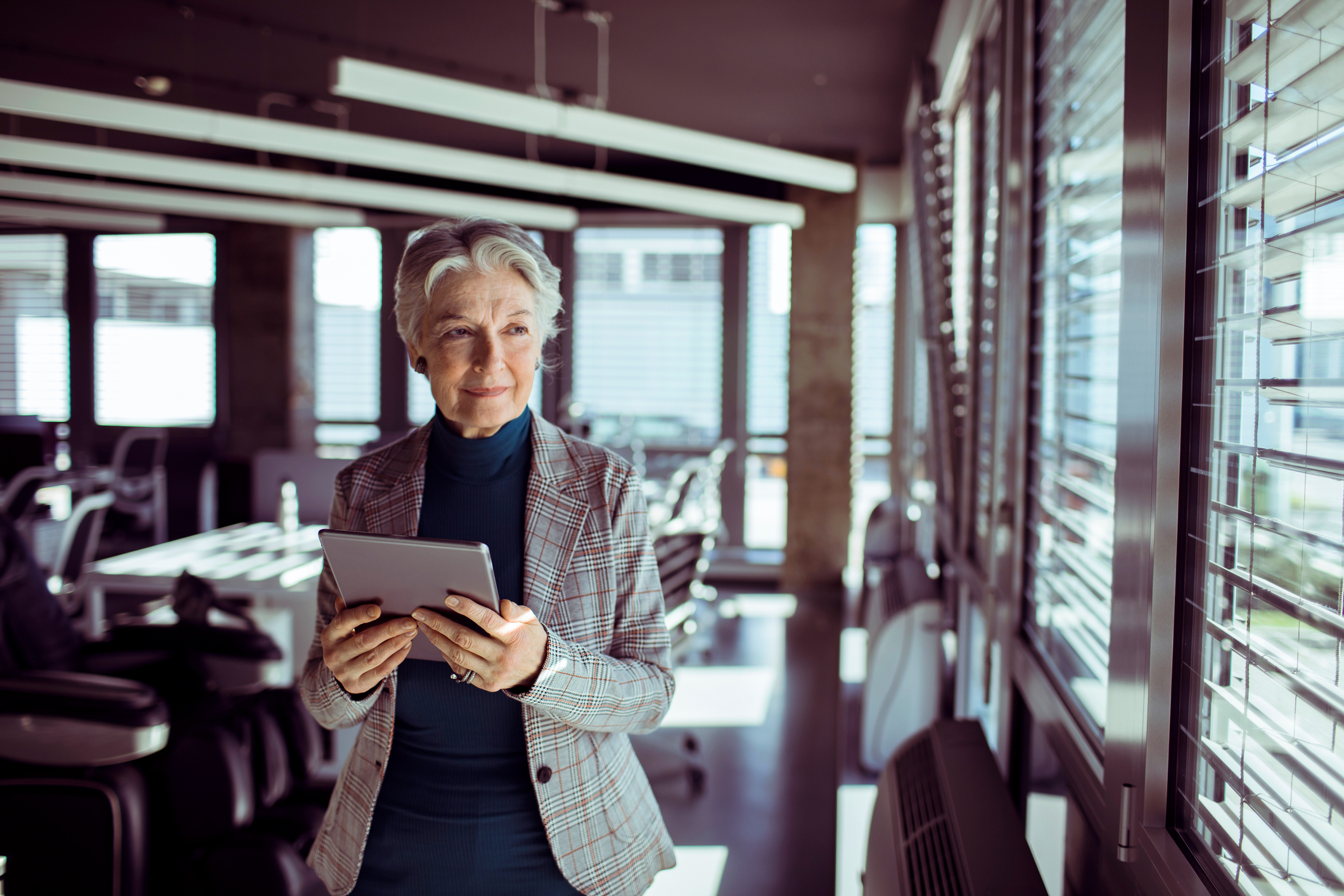 A person standing in a cafe, holding a tablet, looking out of a window with a knowing smile. 