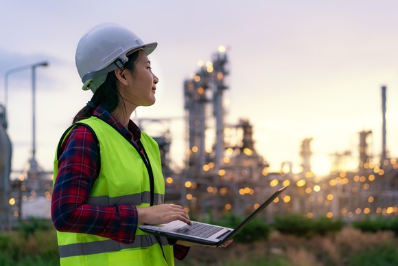 A person in a hardhat holds a laptop near a refinery.