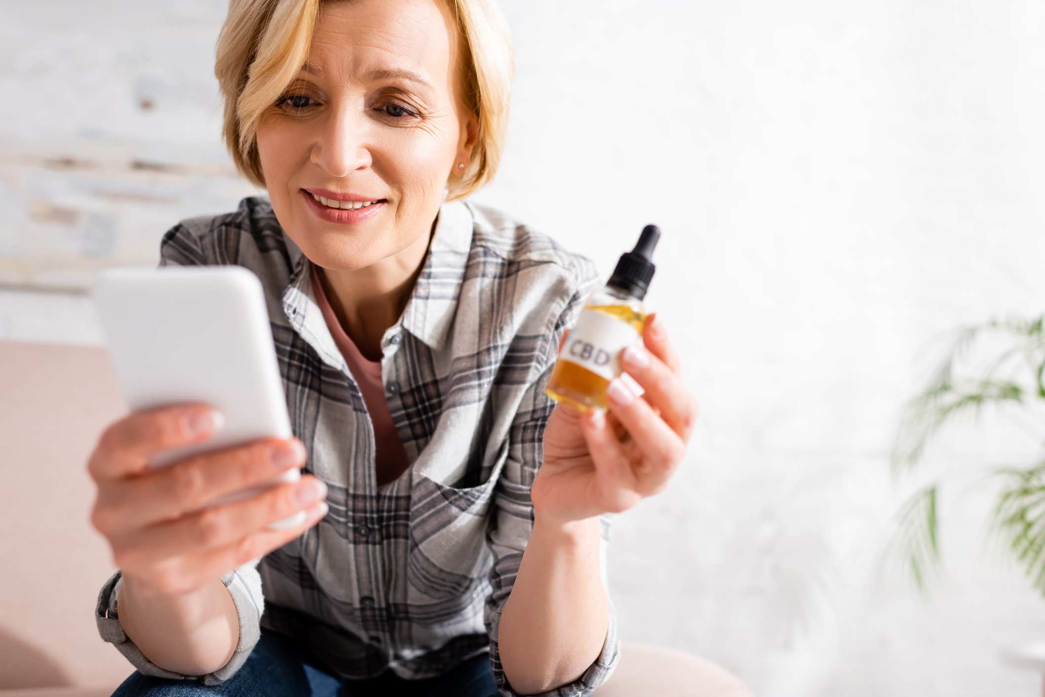 A senior woman holds a bottle of cannabis oil and studies her phone.