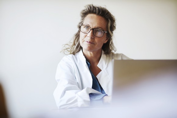 A woman in a lab coat looks up from behind her computer.