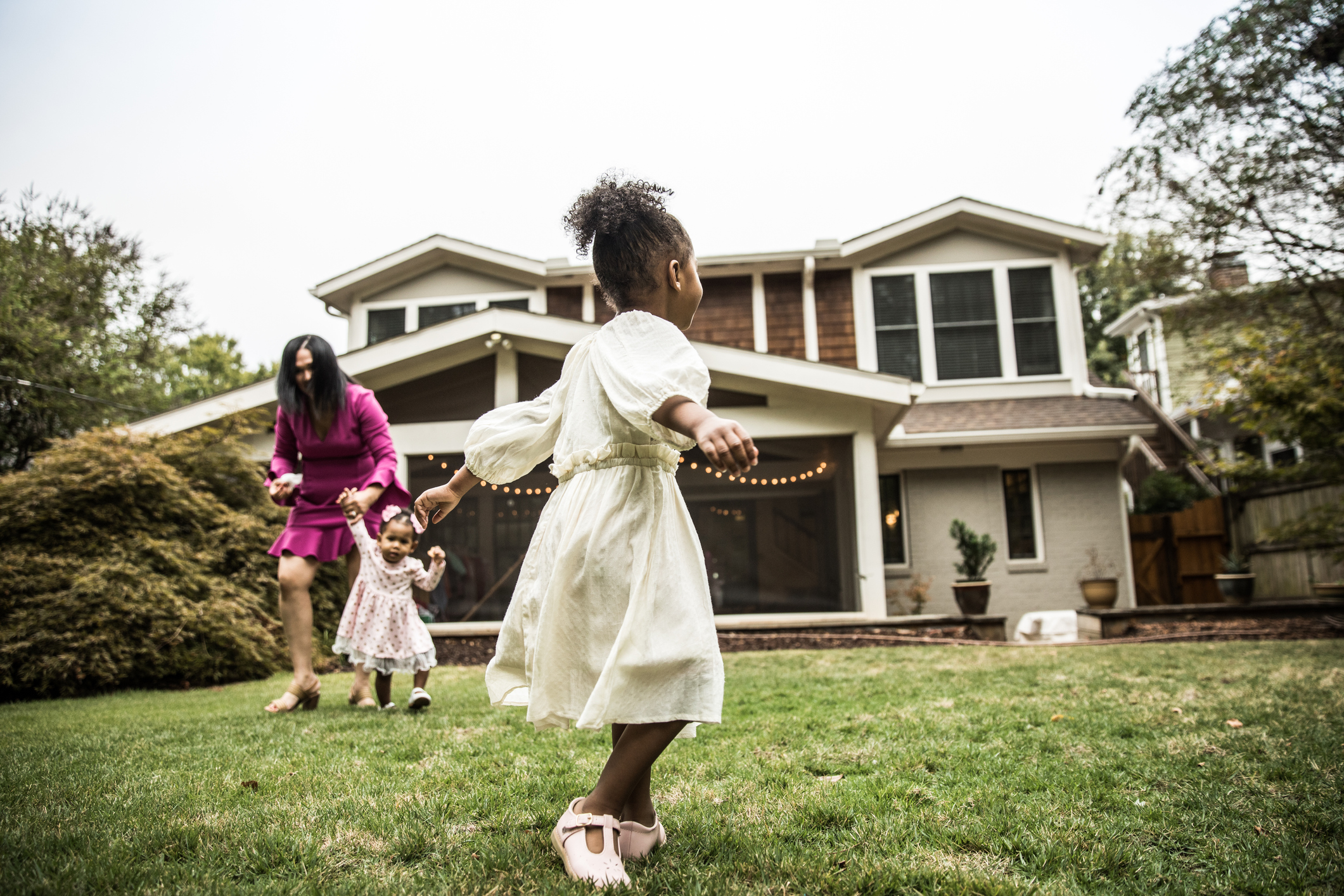 A young child twirling in front of a house with another child and adult on the lawn in the background.