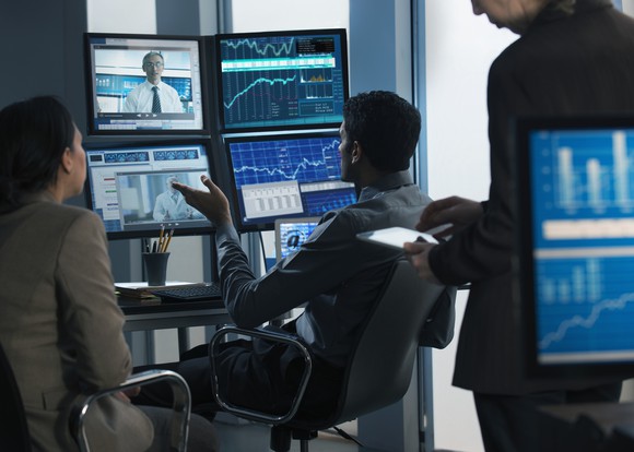 Three people looking at screens showing charts and television programs.