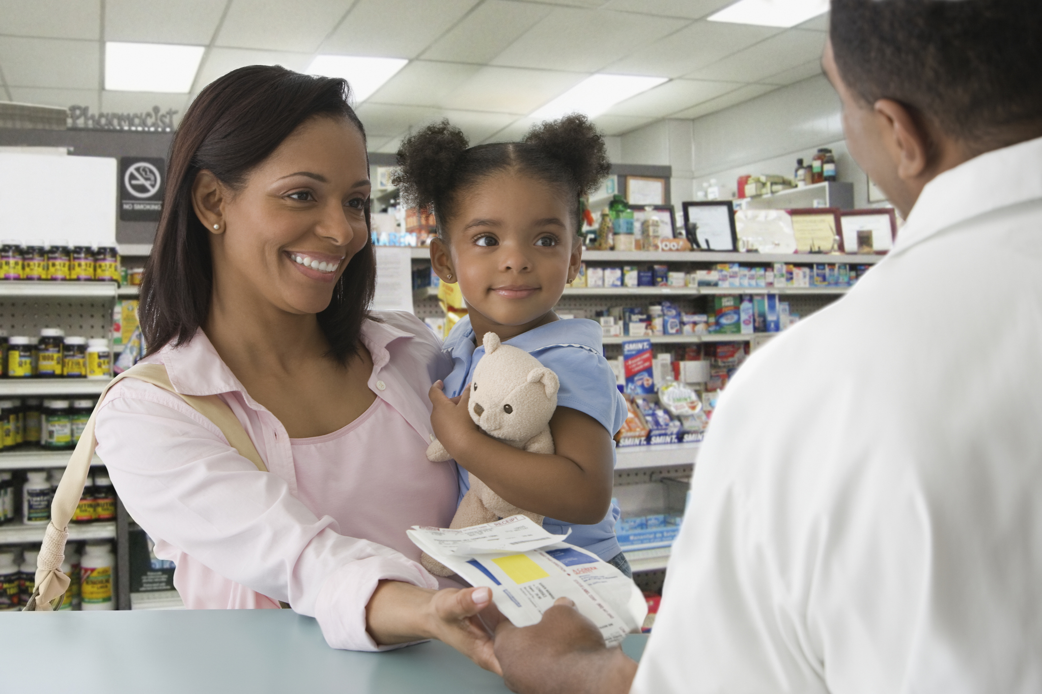 A smiling customer holds a child with a teddy bear at a pharmacy counter.