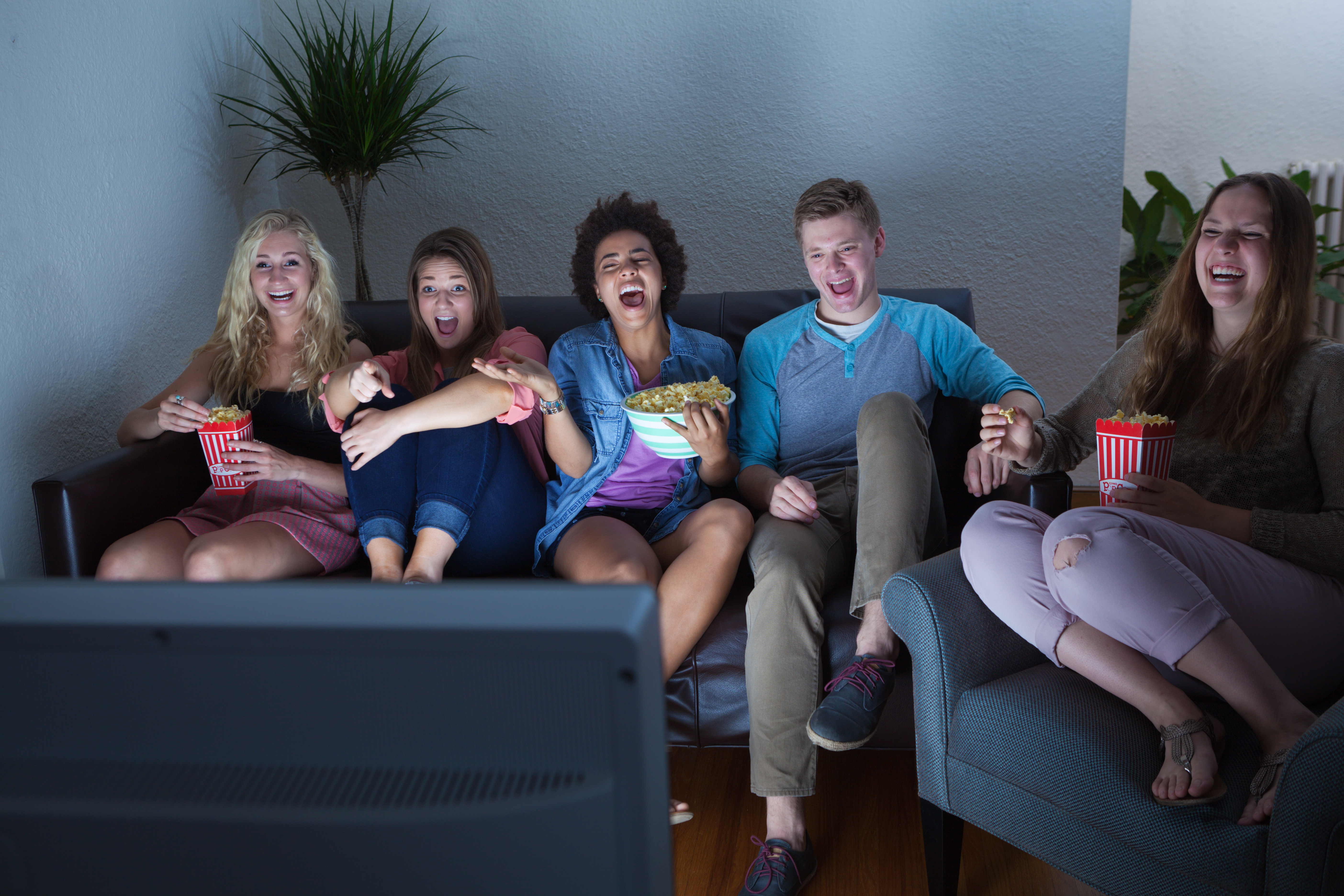 a group of five people sit on a couch and chair in a poorly lit room and laugh at a TV screen in the foreground