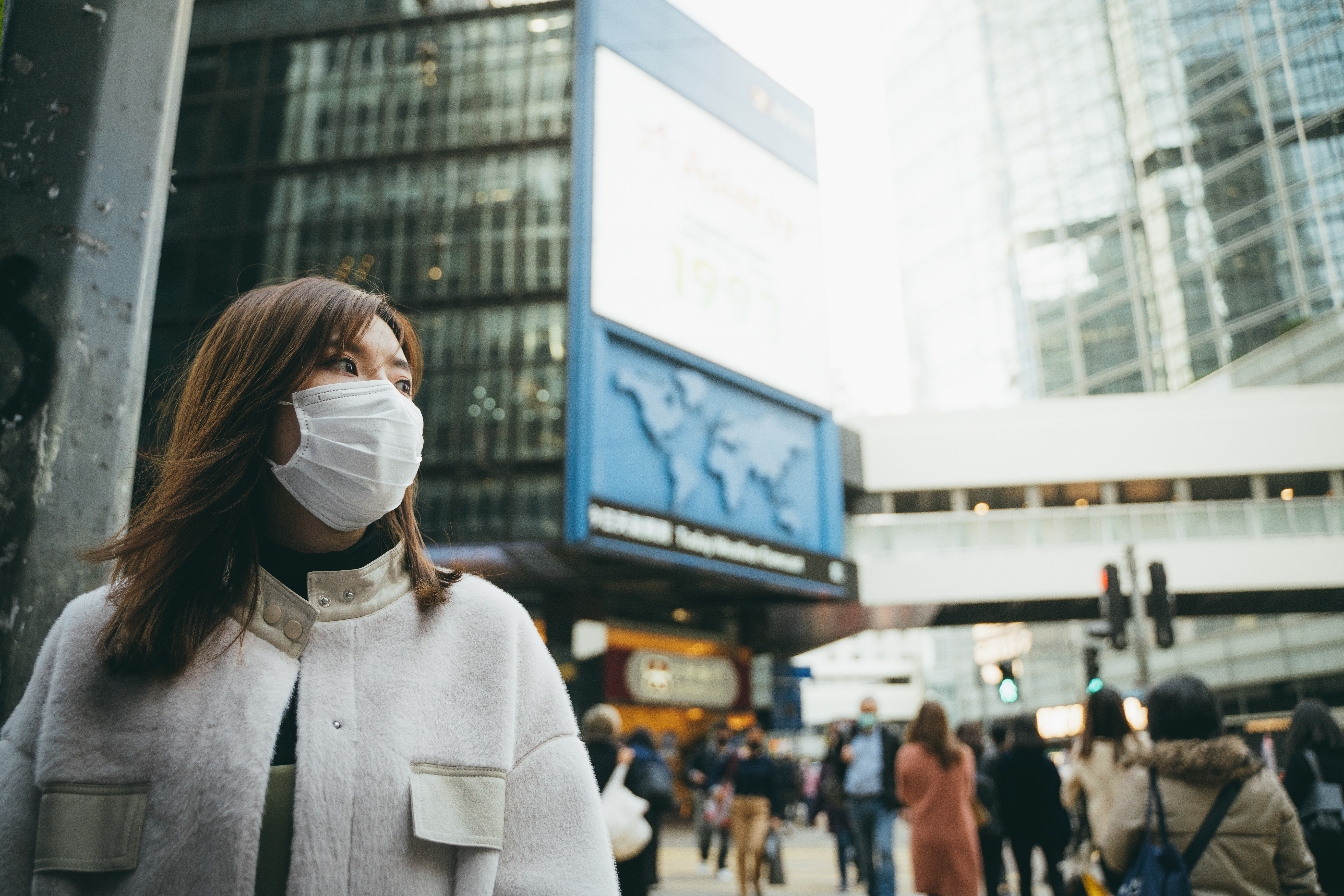 A person wearing a mask and jacket stands outside in a crowd of bustling people.