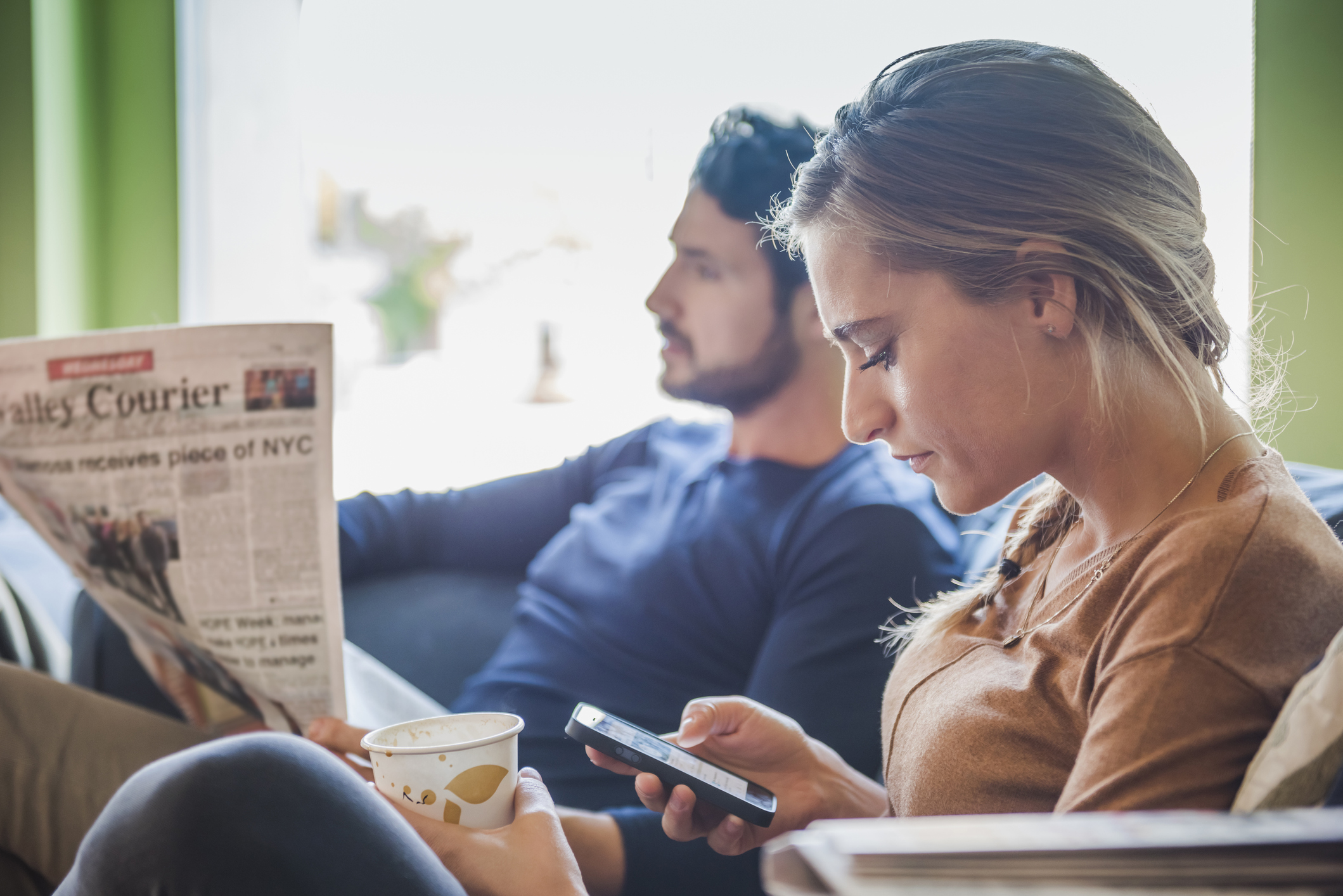 Two people, one of which is reading a newspaper, the other looking at a smartphone.