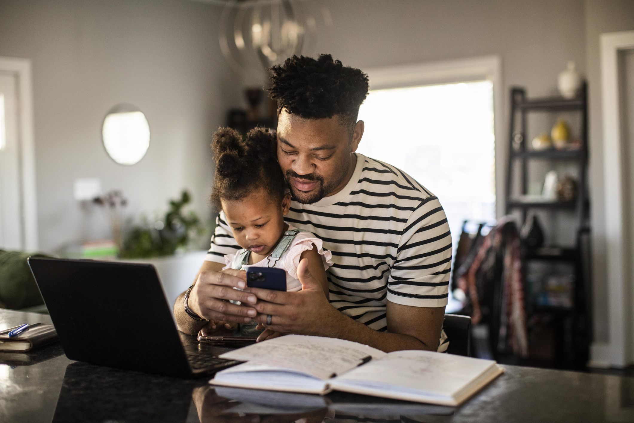 Adult sits at a countertop while scrolling on a smartphone with a small child in their lap.