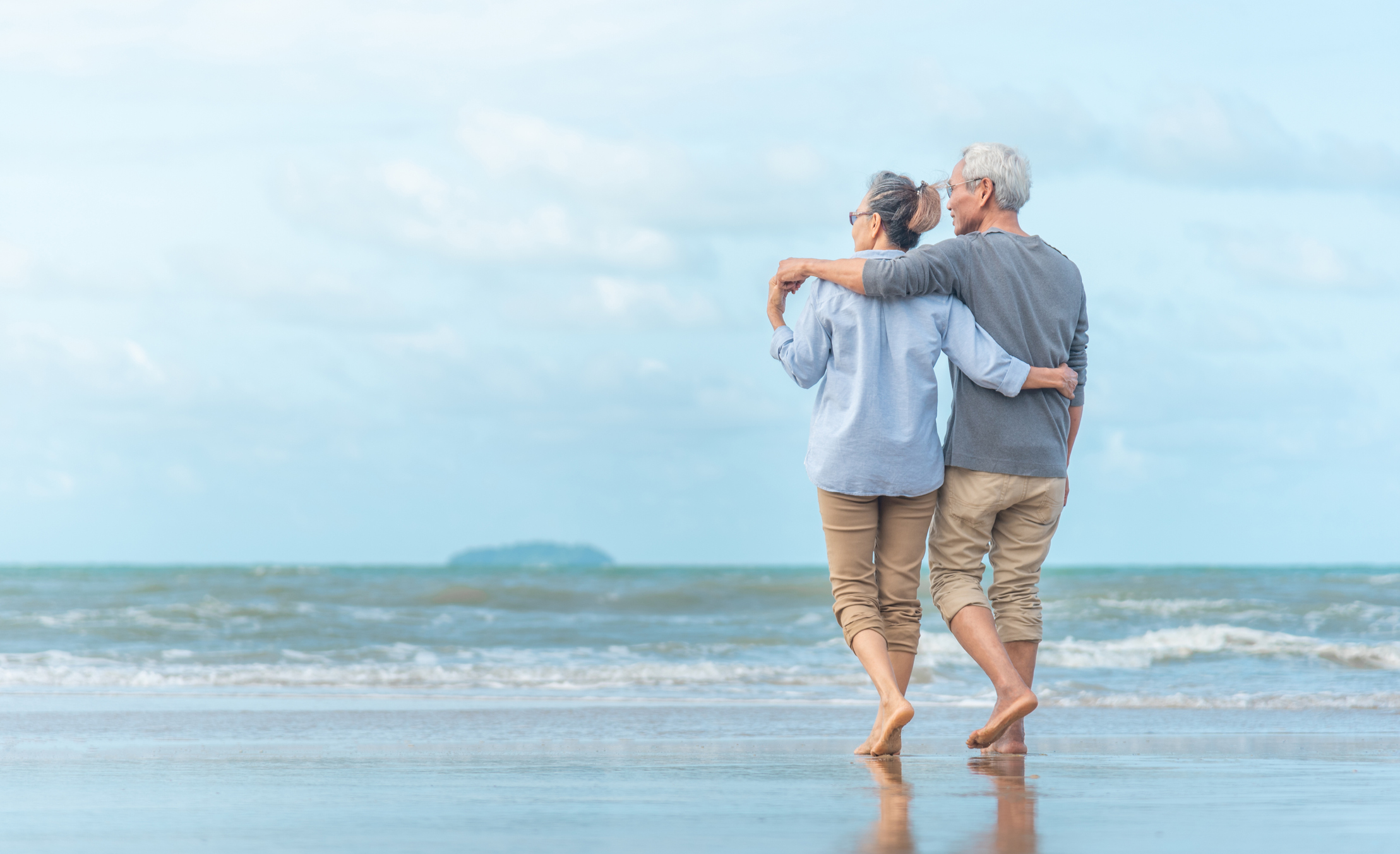 Senior couple walking on the beach with their arms around each other.