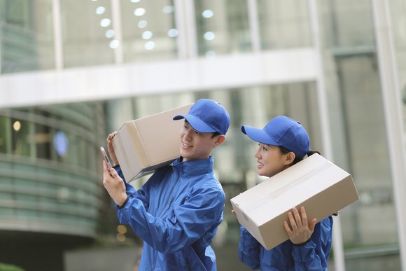 Two delivery people holding cardboard boxes check a smartphone.