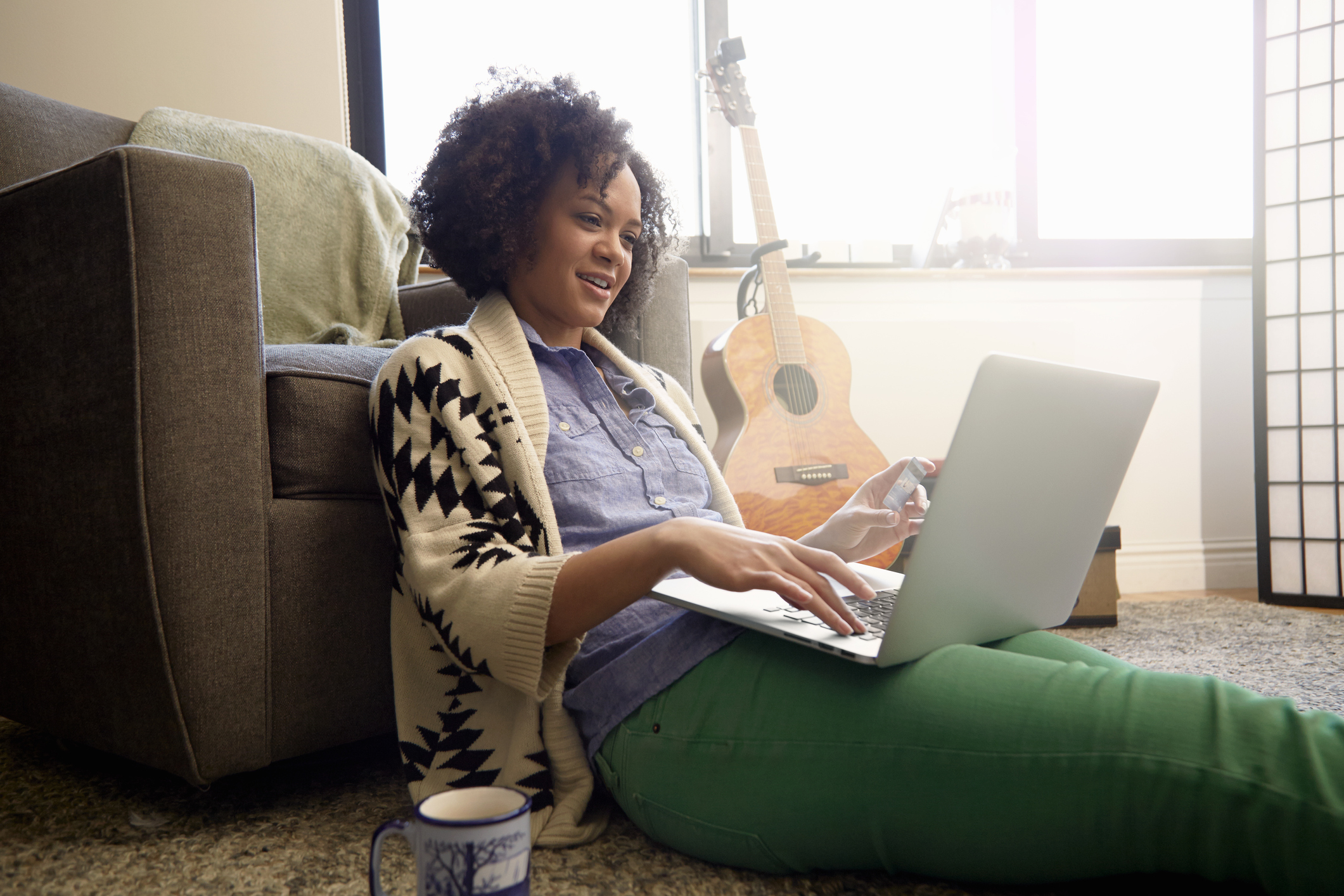 Young adult browses on a laptop while sitting on the floor beside a guitar and mug.