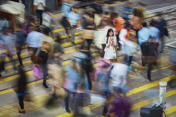 Person standing still and looking at their smartphone while surrounded by blurry images of other people crossing the street.