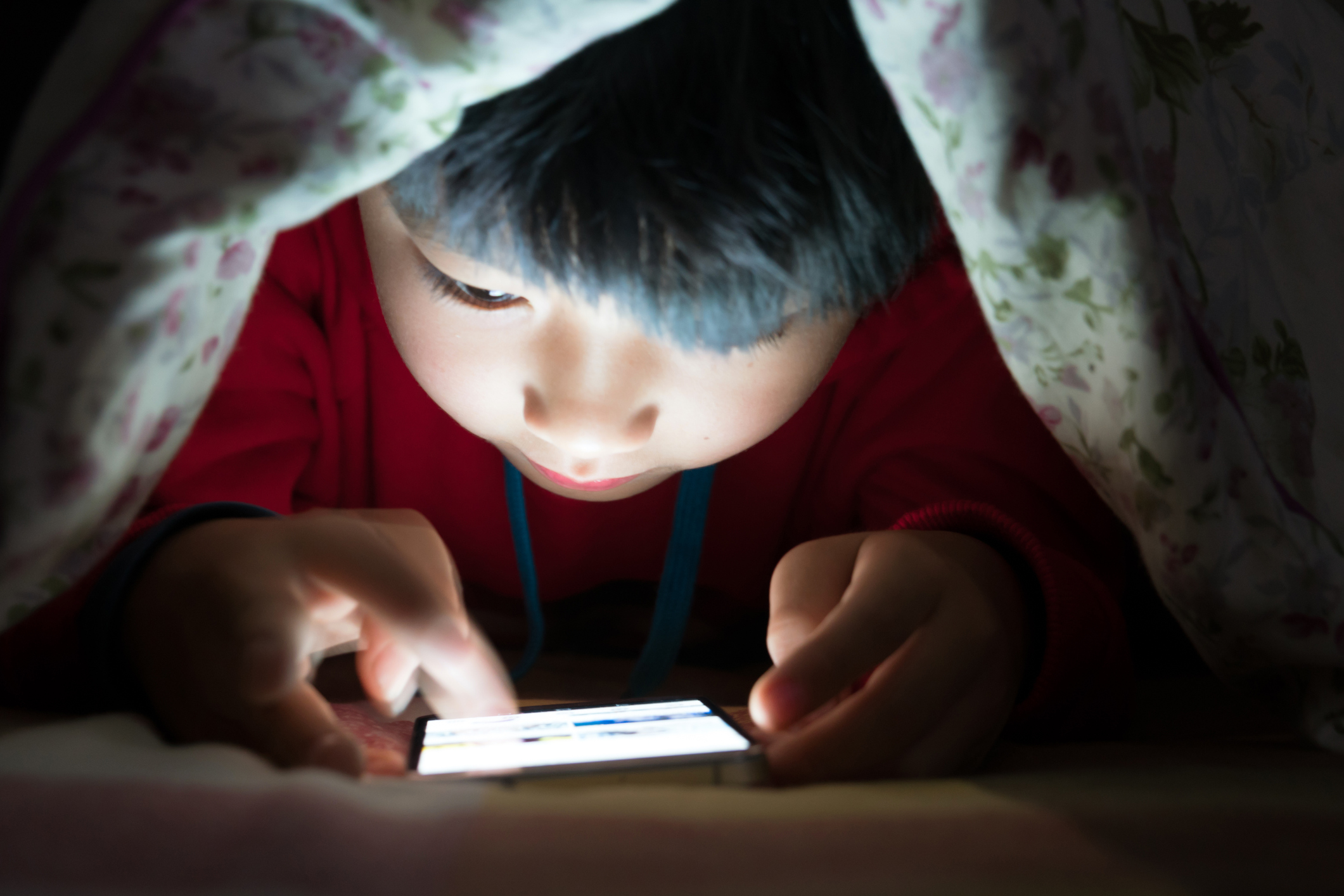 A young boy looks at a smartphone under his covers.
