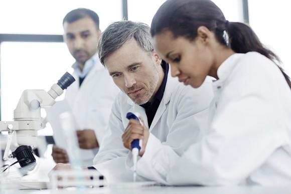 A scientist works at a lab bench while two other scientists look on.