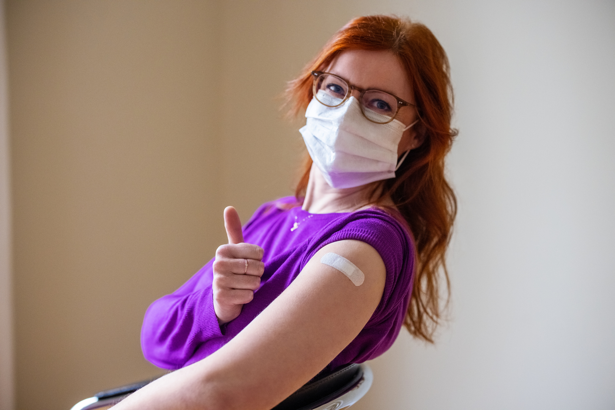 A person in a mask with a bandage on their arm gives a thumbs up to the camera.