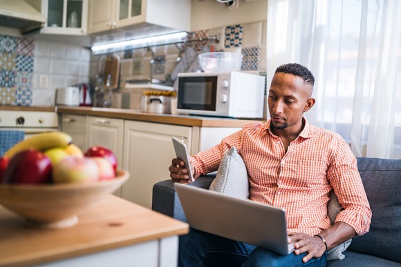 Person holding laptop and smartphone while seated near a kitchen.