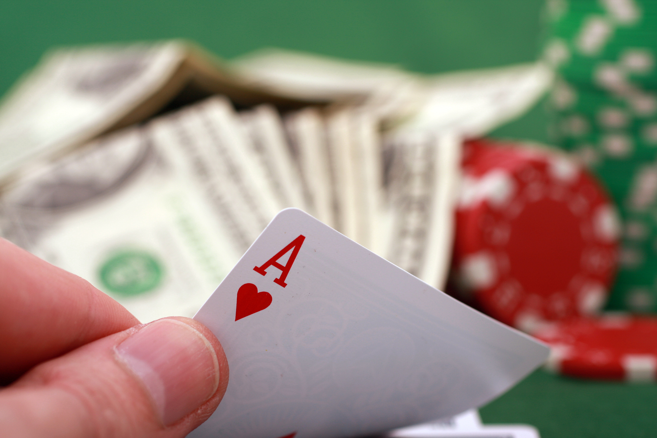 Fingers holding an ace of hearts card, with cash and poker chips in the background.