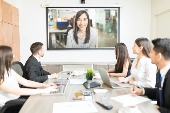 A boardroom of people videoconferencing with a woman on a TV.