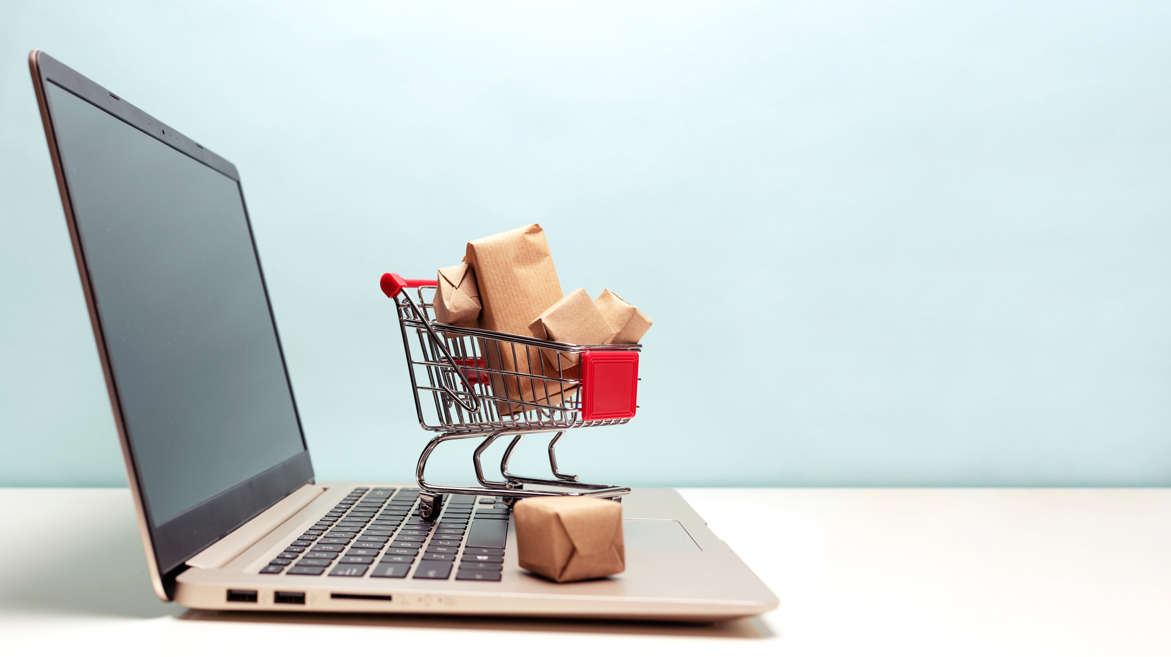 A tiny shopping cart, full of goods and packages, stands on the keyboard of a laptop computer.