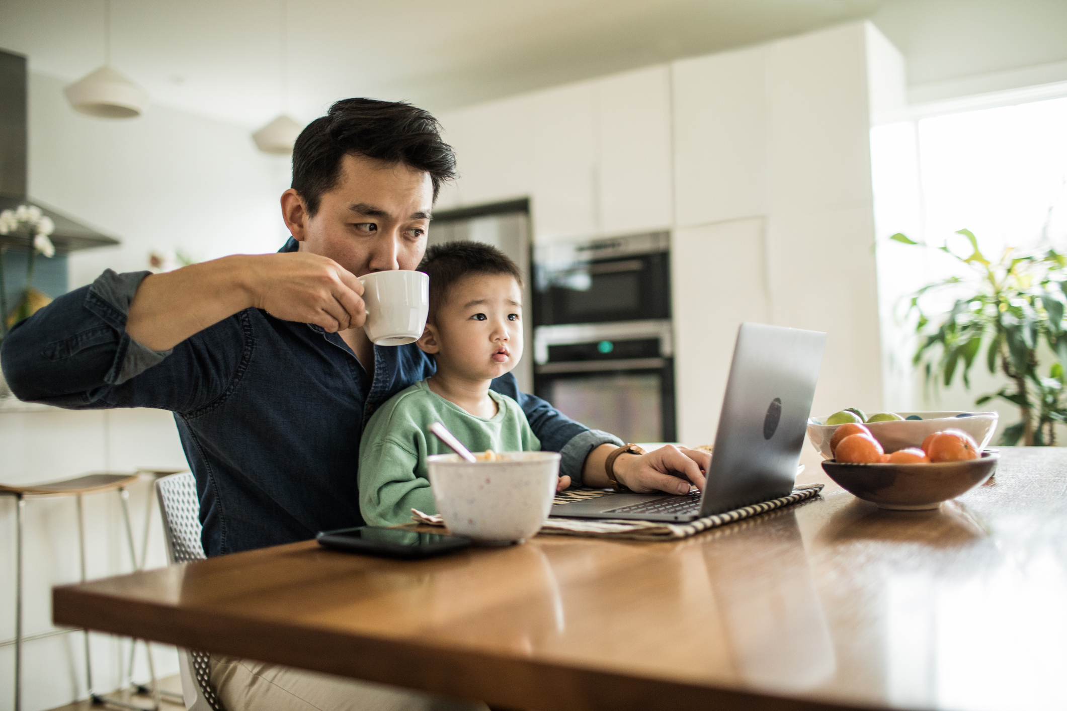 Father works on laptop at breakfast table with young son on lap.