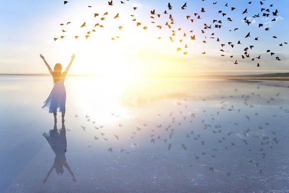A lady lifts her arms while looking over a body of water as a flock of birds glide past during sunrise.
