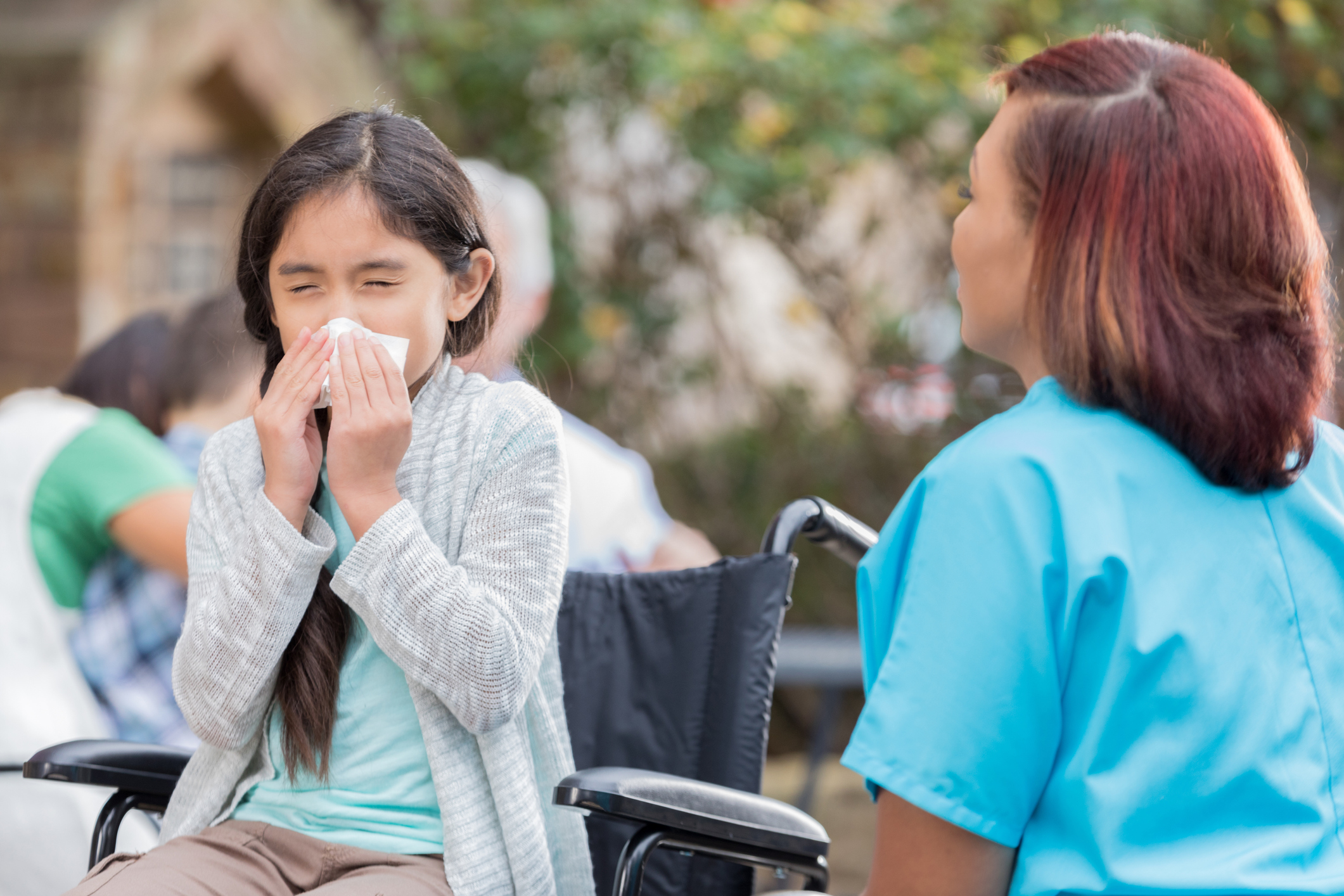 A child sitting in a wheelchair blowing their nose with a tissue as a nurse sits by.