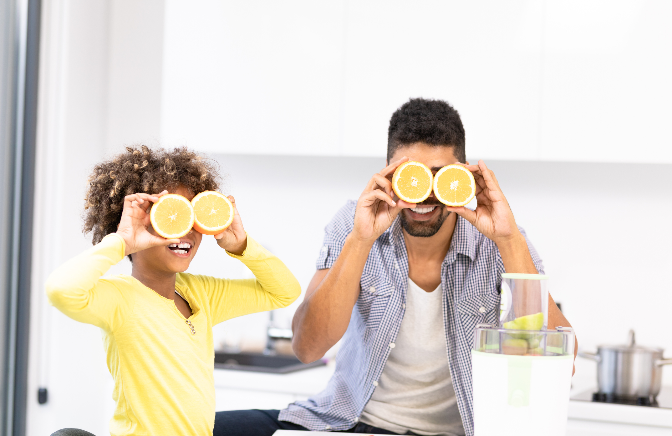 A man and a child in a kitchen holding cut lemons over their eyes.